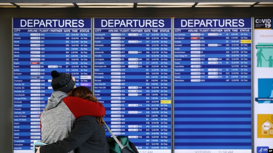 Un hombre y una mujer se abrazan frente a la lista de salidas de vuelos en el Aeropuerto Internacional Dulles, Virginia, el martes 17 de marzo de 2020. Al menos 15 periodistas de los diferentes servicios de idiomas de la VOA que regresarán a su país