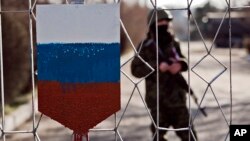 FILE - The former emblem of the Ukrainian army is covered in paint in the colors of Russia's flag as a masked soldier is seen behind a gate to a military base in Simferopol, Crimea, March 13, 2014.